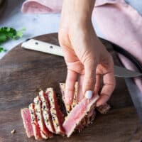 A hand thinly slicing the seared tuna steaks and holding one slice showing the pink inside of the fish and the golden seared sesame crust