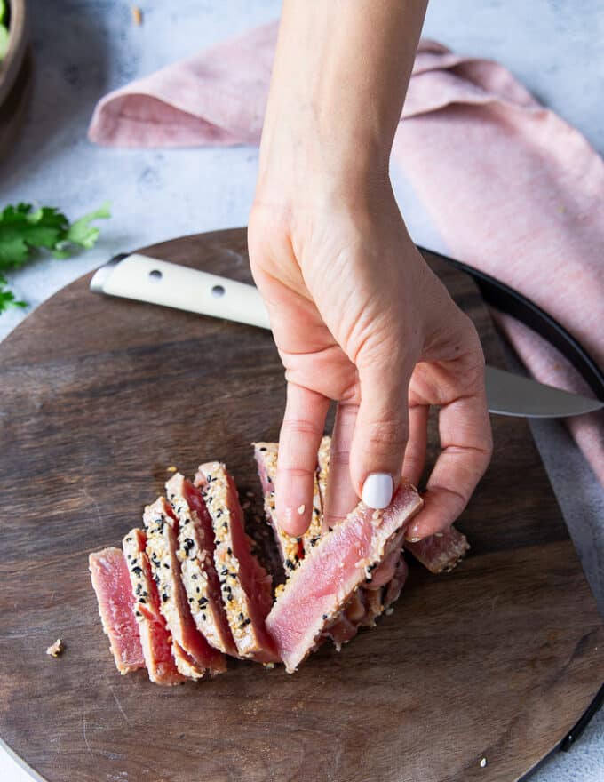 A hand thinly slicing the seared tuna steaks and holding one slice showing the pink inside of the fish and the golden seared sesame crust