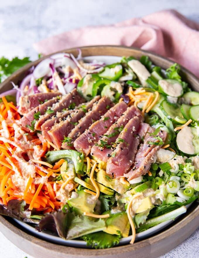 close up of a sliced ahi tuna salad showing the pink inside of the ahi tuna and the sesame crust on the outside, drizzled with a goma sesame dressing on a bed of avocado, cucumber, carrots, crispy wontons and more