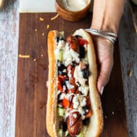An assembled Mediterranean style hot dog showing the grilled hot dog with tahini, diced tomatoes and cucumbers , black olives, feta cheese