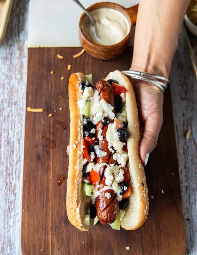 An assembled Mediterranean style hot dog showing the grilled hot dog with tahini, diced tomatoes and cucumbers , black olives, feta cheese