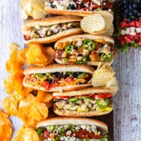 A board with 6 different assembled grilled dogs from the hot dog bar showing the variety of toppings. Some chips and a watermelon salad surrounding the board