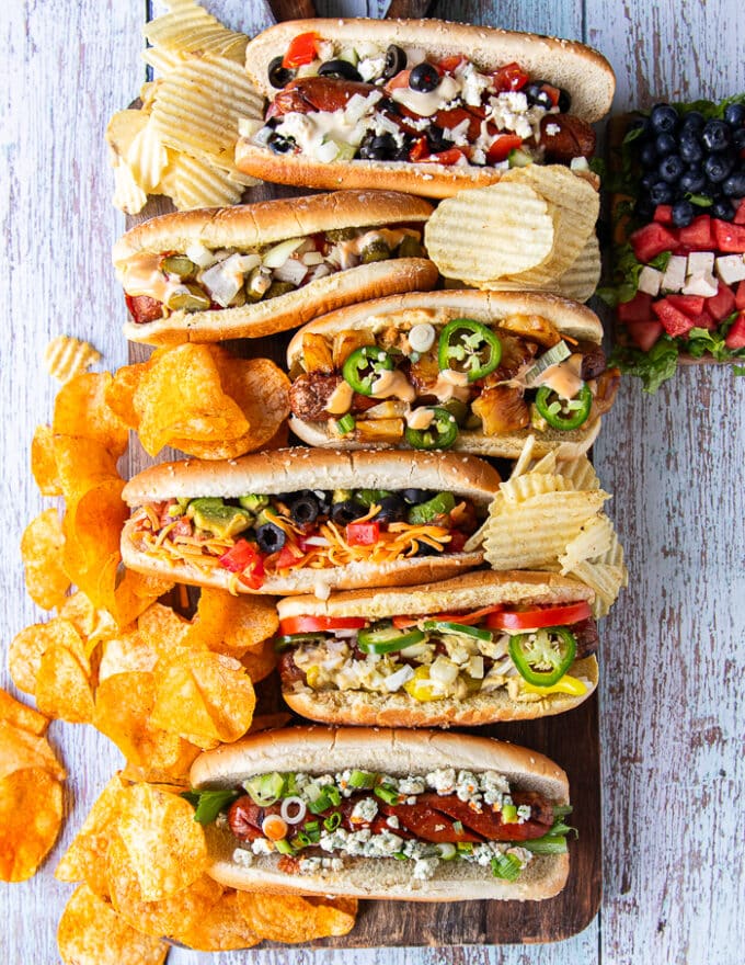 A board with 6 different assembled grilled dogs from the hot dog bar showing the variety of toppings. Some chips and a watermelon salad surrounding the board 