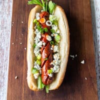 An assembled buffalo style hot dog showing the grilled hot dog with blue cheese, celery, hot sauce and green onions