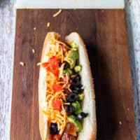 An assembled Nacho style hot dog showing the grilled hot dog with salsa, guacamole, cheddar cheese, green onions, black olives, diced tomatoes and onions