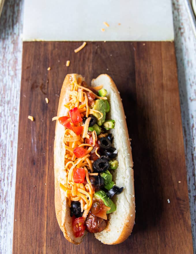 An assembled Nacho style hot dog showing the grilled hot dog with salsa, guacamole, cheddar cheese, green onions, black olives, diced tomatoes and onions