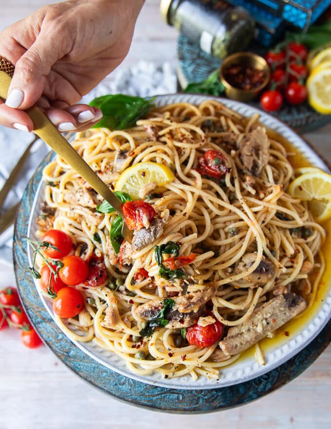 A hand holding a fork on a plate of sardine pasta showing the sardines, basil, tomatoes, capers, lemon and olive oil sauce around it 