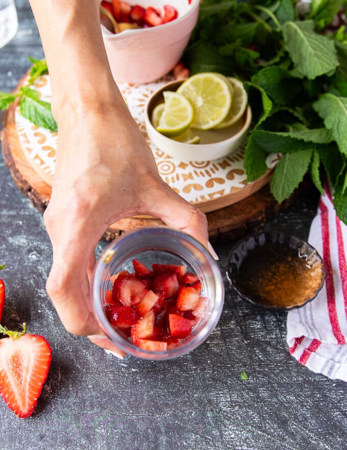 A hand holding a cup with diced strawberries to make strawberry mojito