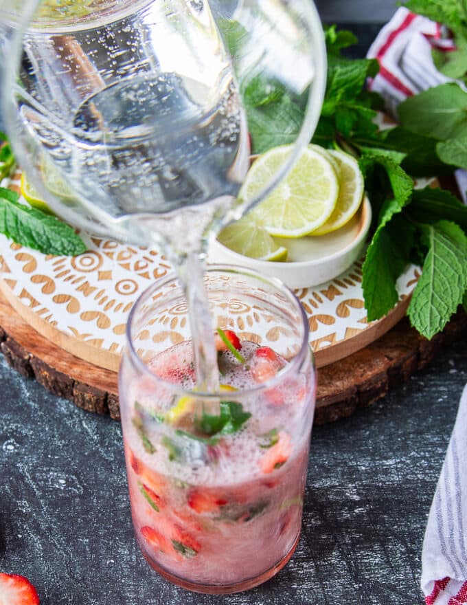 Club soda is poured over the muddled fruit and mint showing some fizz in the cup and continuing to pour until it fills up the cup.