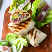 A cutting board with tuna wraps, some of them cut up to show the filling and the rest are not. Tuna lettuce wraps are also assembled and placed next to the regular tuna wrap recipe on the cutting board