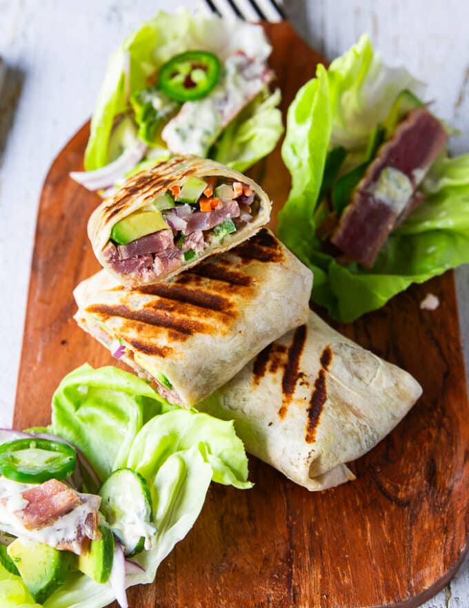 A cutting board with tuna wraps, some of them cut up to show the filling and the rest are not. Tuna lettuce wraps are also assembled and placed next to the regular tuna wrap recipe on the cutting board 