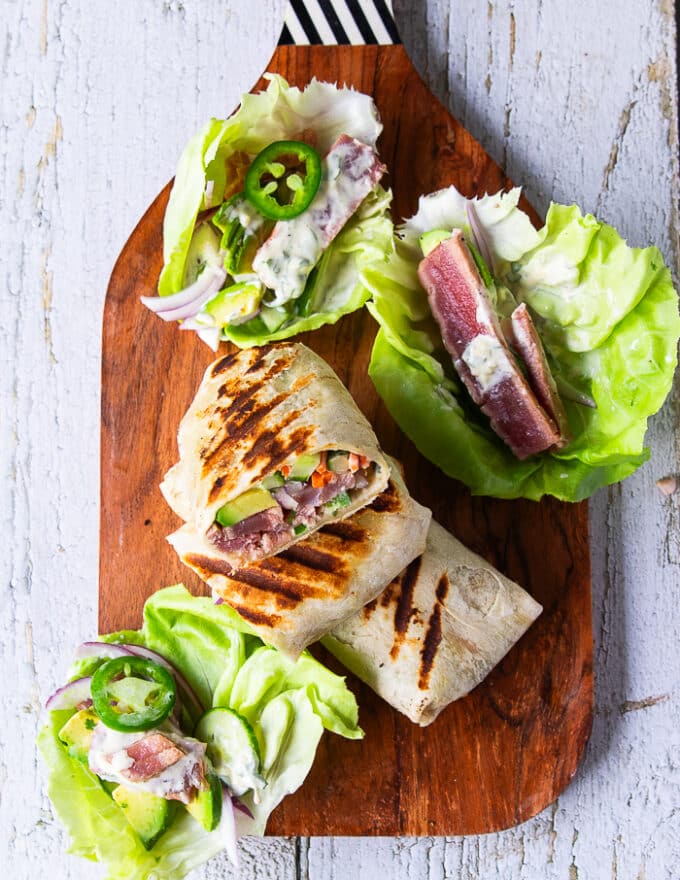 Tuna wraps on a cutting board with one cut in half showing the tuna and filling, and some tuna lettuce wraps assembled ad ready as well served for a crowd 
